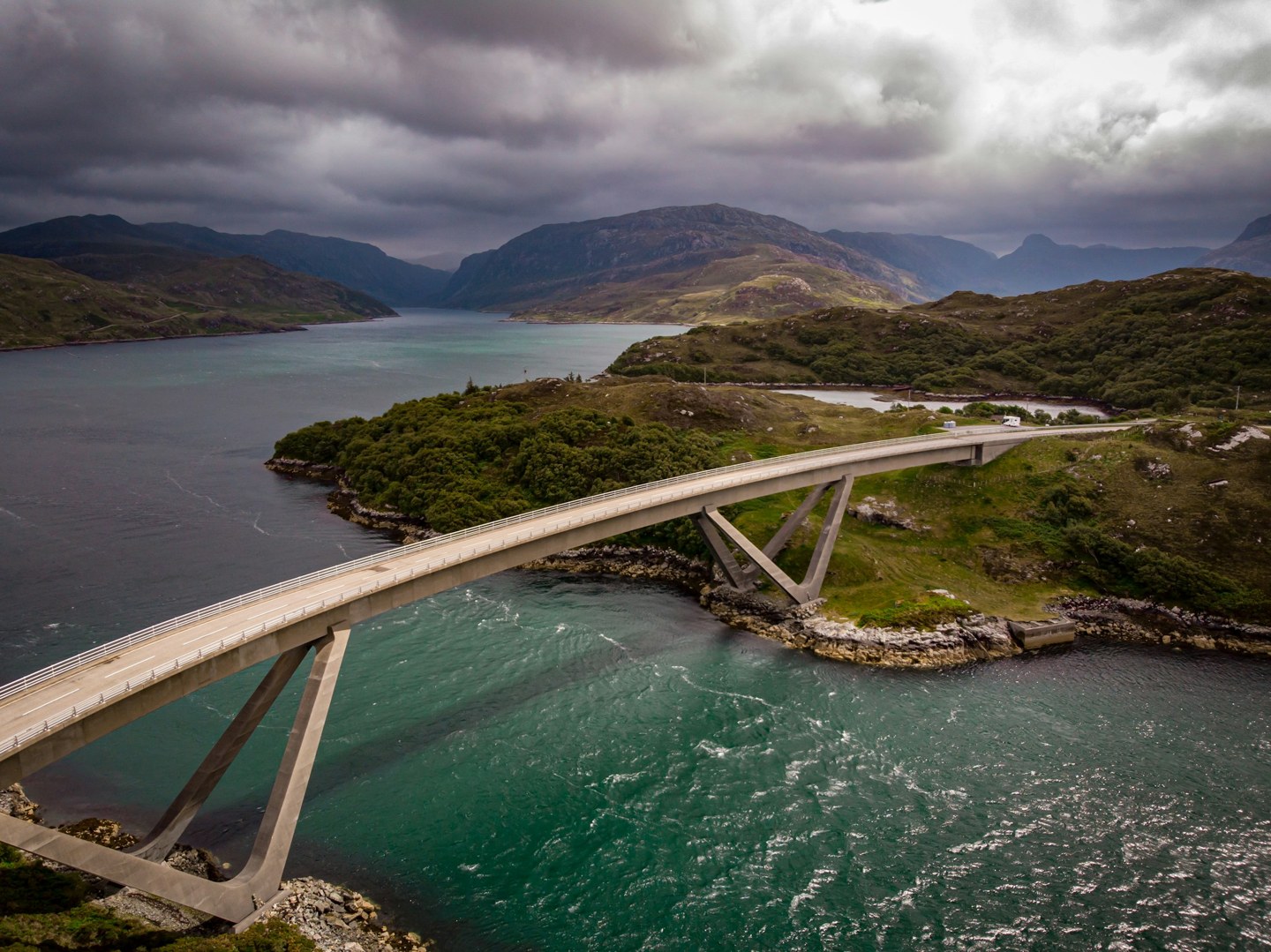 Aerial view of Kylesku Bridge curving across water between mountains in the Scottish Highlands