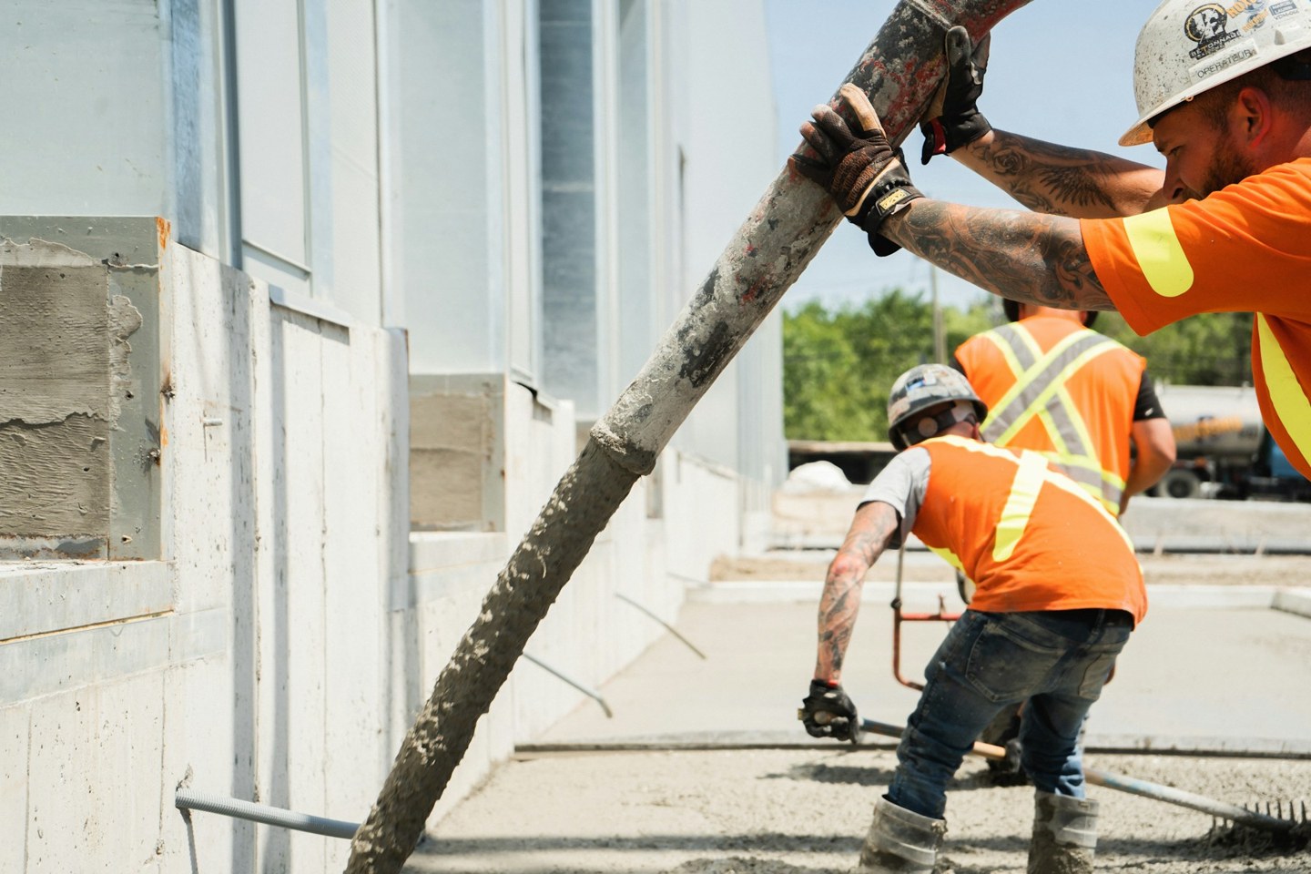 Construction workers guiding a concrete pump hose and levelling fresh concrete beside a building