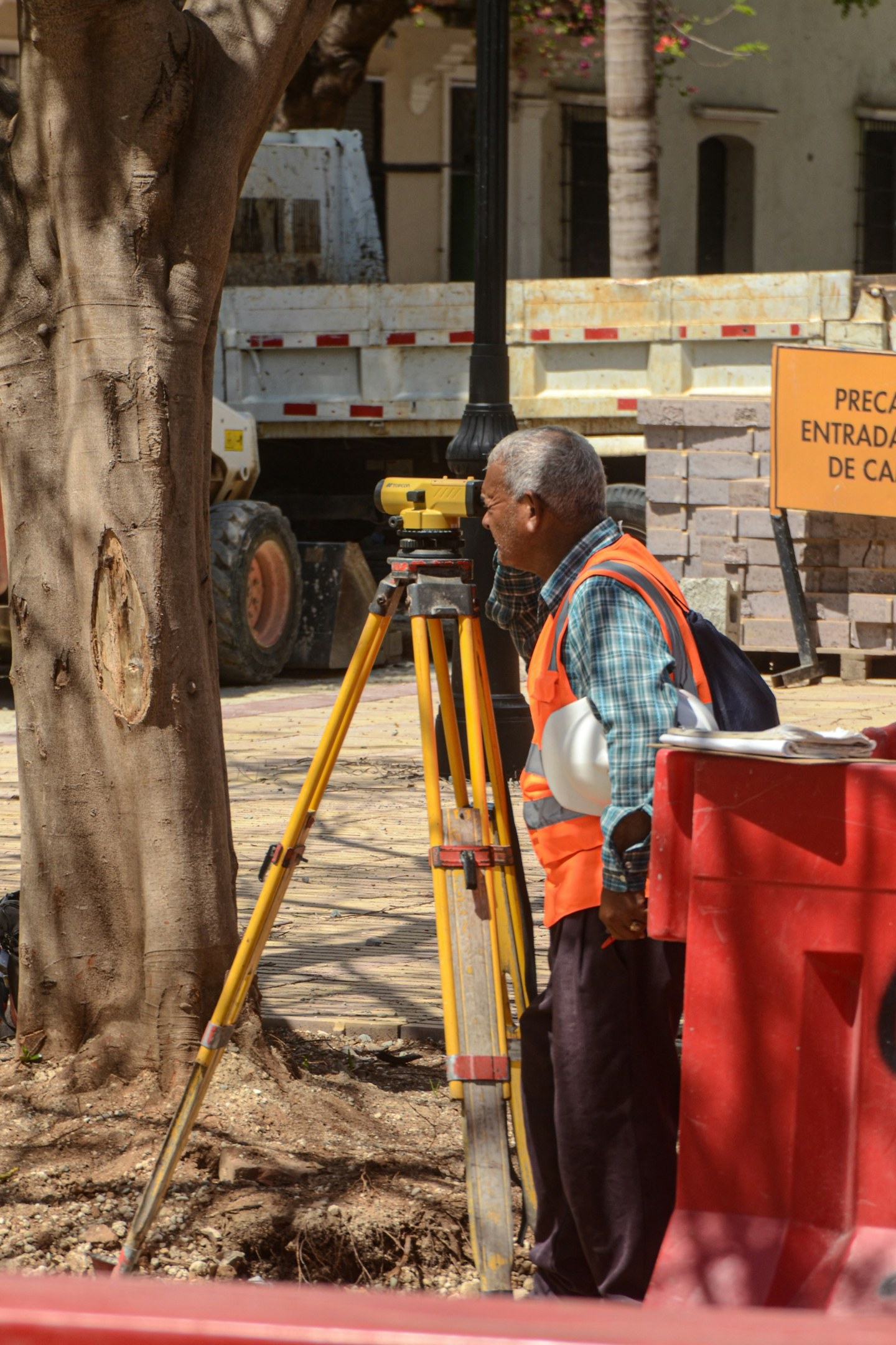 A surveyor in an orange safety vest using a tripod-mounted optical instrument at a street works site