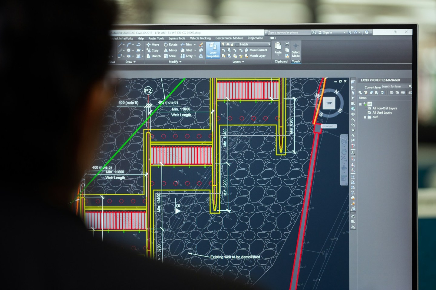 A civil engineer working at a monitor displaying a detailed engineering drawing in CAD software