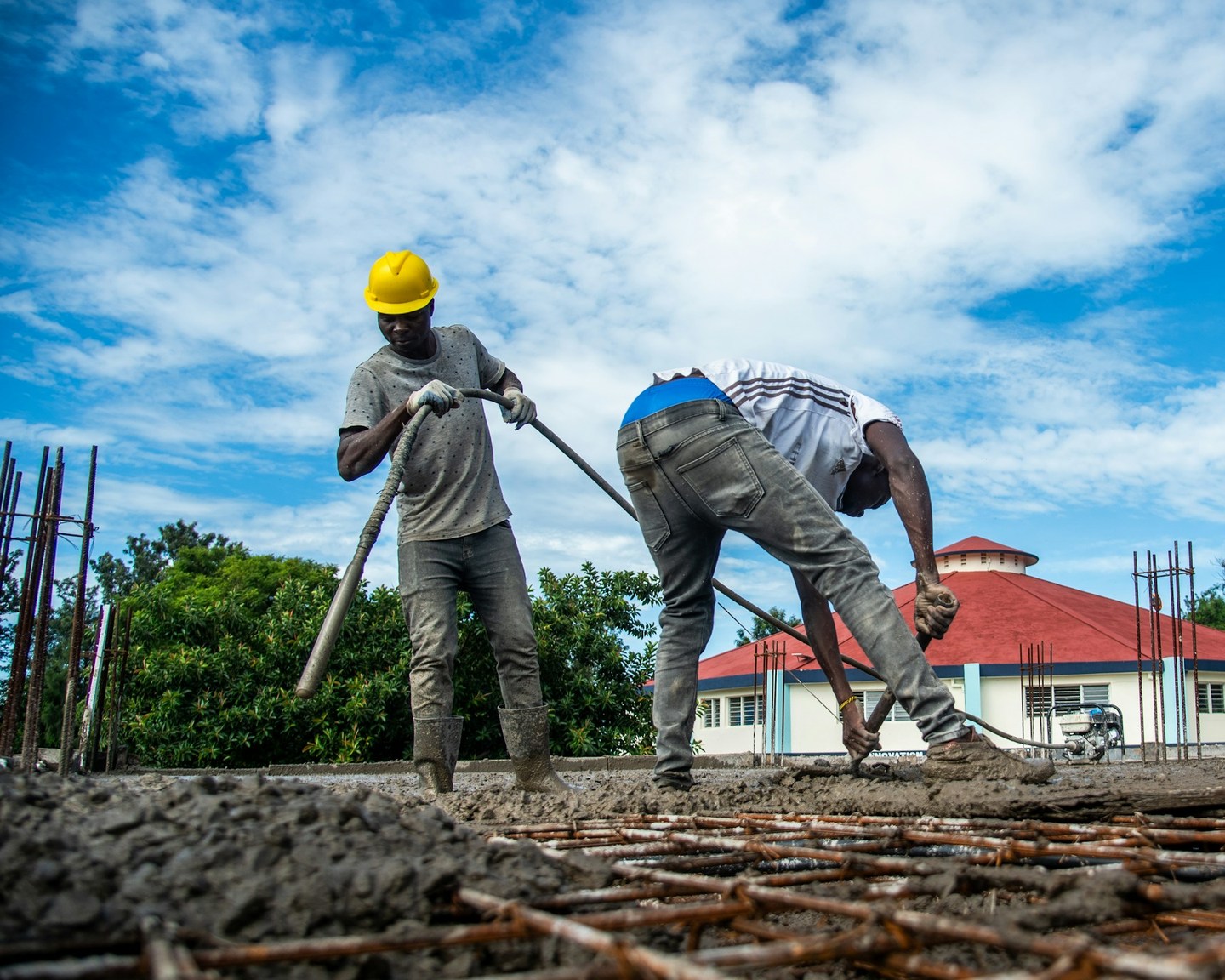 Two workers spread fresh concrete over a reinforcement grid beneath a blue sky