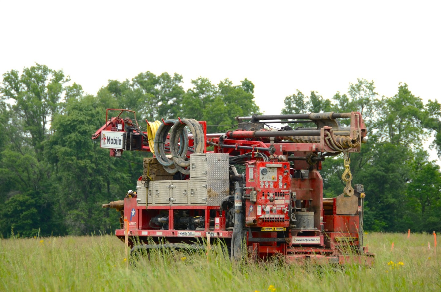 A red tracked mobile drilling rig parked in a grassy field with trees behind