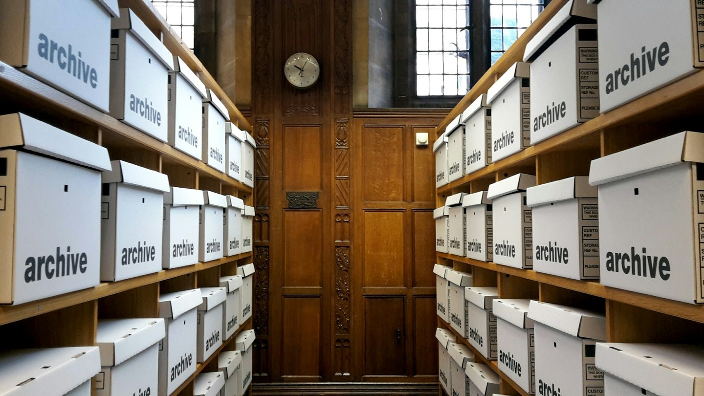 An aisle between wooden shelves holding white archival storage boxes