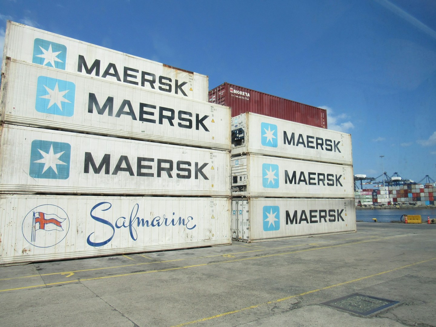 Shipping containers stacked on a paved terminal with cranes visible beyond