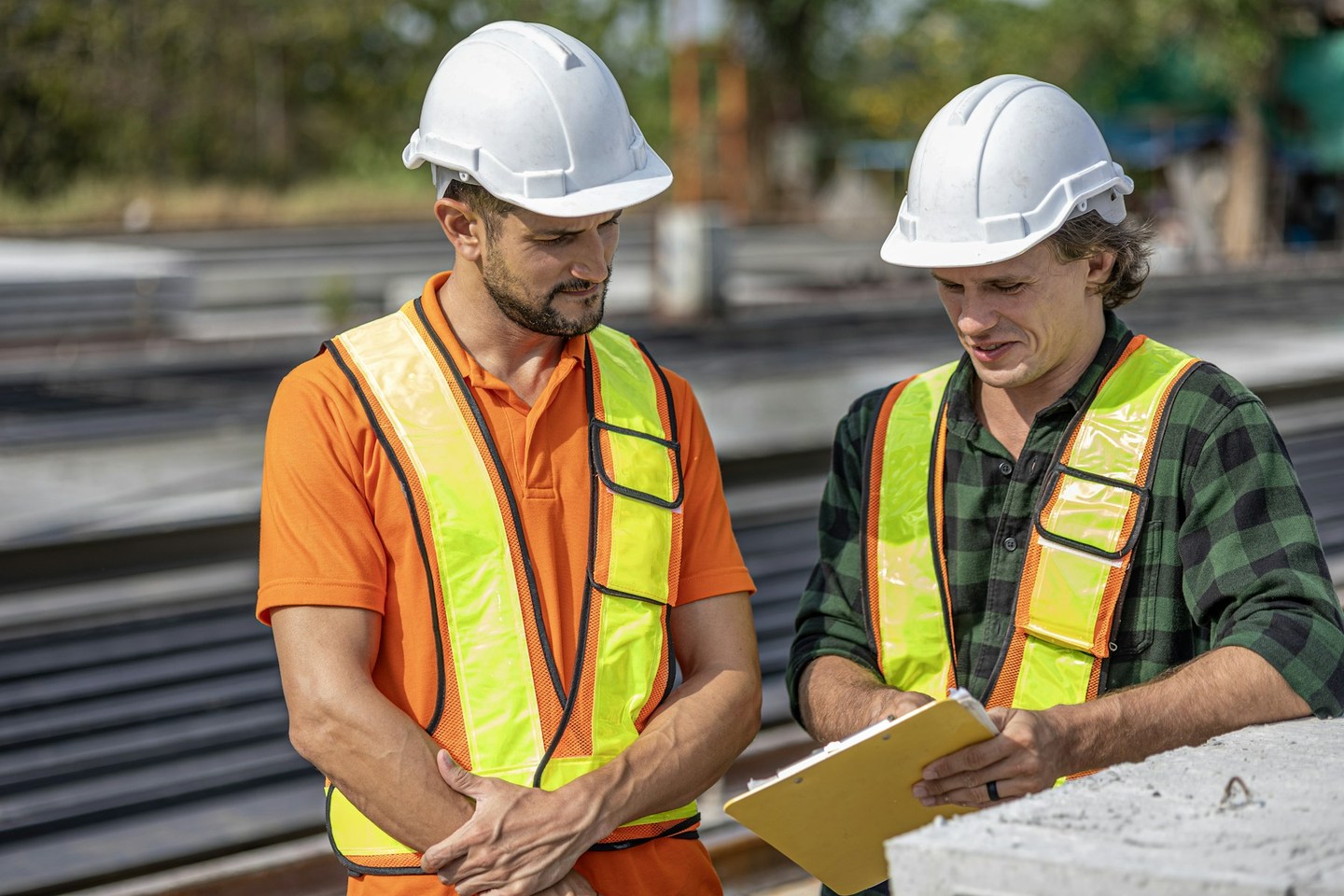 Two people wearing hard hats and high-visibility vests review a clipboard beside precast concrete